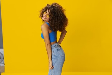 Woman with curly afro hair in a blue T-shirt on a yellow background dancing flying hair with sunglasses yellow, hand signs, look into the camera, smile with teeth and happiness, copy space