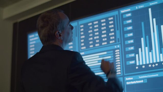 Businessman analyzing data and statistics shown on big screen. Back view of male manager in official suit in dark monitoring room working on project and processing information. Technology concept - Powered by Adobe