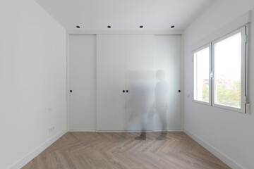 Built-in wardrobe with white wooden sliding doors in a room with aluminum windows and a ghost passing in front of the wardrobe