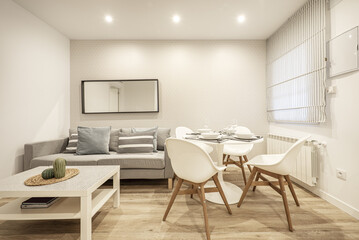 Living-dining room of a house furnished with a gray sofa, a window with a fabric blind and a round dining table