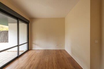 Small empty room with brown wooden flooring with cream walls and a large window with sliding doors leading to a patio