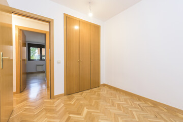 Empty room with herringbone oak parquet flooring throughout the house, built-in wardrobe with three oak doors and access door of the same material