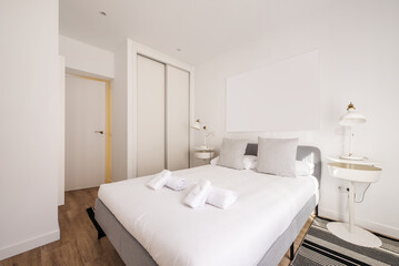 Fototapeta na wymiar Bedroom with a gray fabric double bed and white duvet with clean rolled towels and a built-in wardrobe with white wooden sliding doors