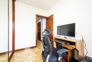Bedroom without bed with wooden desk with swivel chair with large pc screen with back to built-in wardrobe with sliding mirror doors