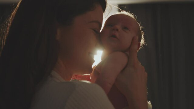 Portrait of smiling young mother with little baby son standing in sunset light shining through window. Concept of family happiness and parenting. Shot in slow-motion