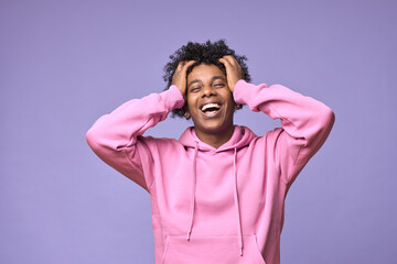 Young cheerful happy hipster African American teen guy wearing pink hoodie isolated on light purple background. Smiling cool ethnic generation z teenager model laughing standing holding head in hands.