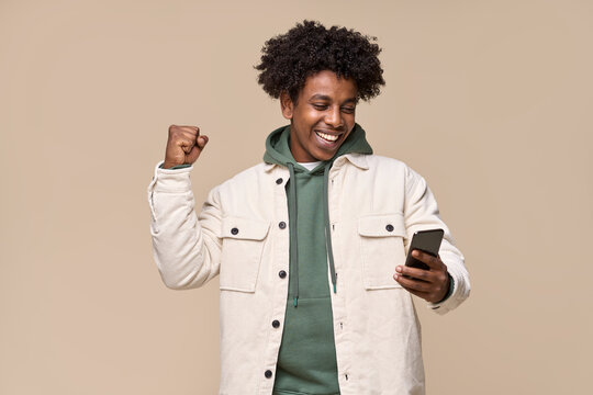 Happy African American Teenager, Ethnic Cool Guy Student Winner Holding Cell Phone Winning Money Online On Cellphone, Using Mobile App Celebrating Great Result With Yes Isolated On Beige Background.