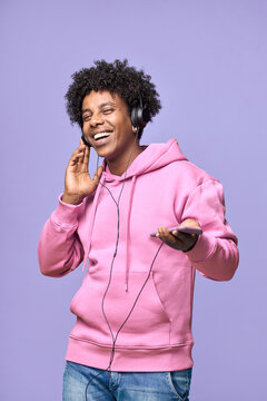 Happy cheerful African American teen boy wearing headphones holding mobile using cellphone dancing listening music on cell phone technology, standing isolated on light purple background. Vertical