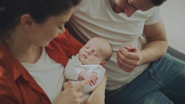 Close-up of a cute newborn baby sleeping peacefully in smiling parent's arms. Husband and wife gently touchng their baby while sitting on sofa