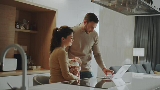 Young Parents In The Kithcen. Mother Sitting With Holding Baby In Arms While Father Standing And Working On Laptop In Slow-motion