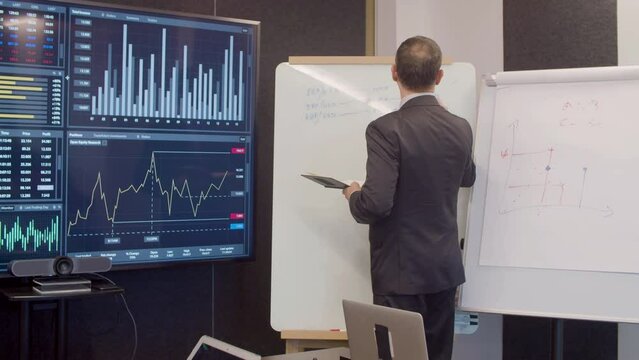 Businessman writing on whiteboard in meeting room. Back view of data analyst looking at digital screen with bar charts and statistics, creating development strategy. Occupation, technology concept
