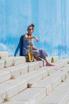 Side view of a woman sitting and leaning on wall, holding a saxophone