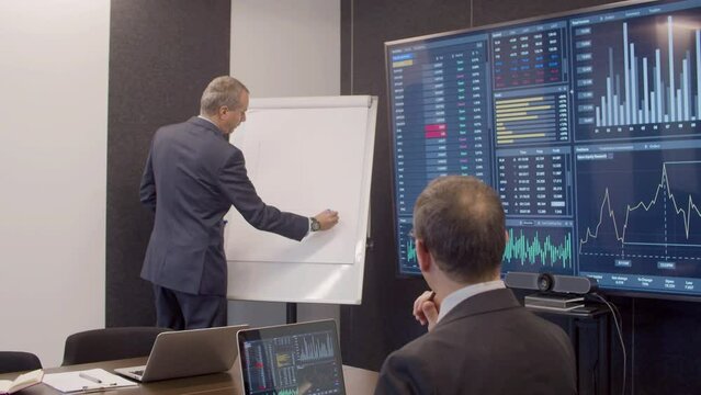 Data engineer drawing on whiteboard in meeting room. Medium shot of man in official suit working with colleague in office with big digital screen, processing information. Teamwork, big data concept