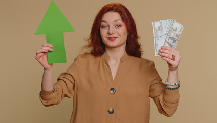 Happy redhead woman winner holding arrow sign pointing up, career growth and money dollar exchange increase. Young ginger girl showing up indicating successful development progress on beige background