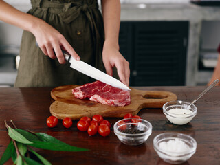 Woman with knife in hand cutting steak meat for frying in kitchen with salt pepper and other spices on table, red cherry tomatoes and herbs, dinner preparation.