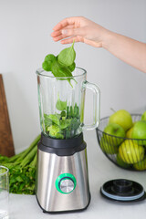 Woman adding spinach leaves into blender with ingredients for smoothie at table indoors, closeup