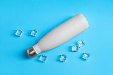Stylish thermo bottle with water drops and ice cubes on light blue background, flat lay