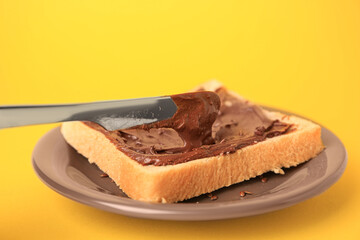 Spreading chocolate paste onto bread on yellow background, closeup