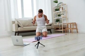 Fototapeta na wymiar Man sports, watching a workout tape on his phone and repeating exercises sports blogger with dumbbells, pumped up man fitness trainer works out at home, the concept of health and body beauty