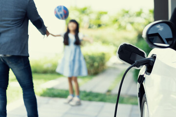 Fototapeta Progressive father and daughter plugs EV charger from home charging station to electric vehicle. Future eco-friendly car with EV cars powered by renewable source of clean energy. obraz