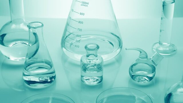 Background shot for skin moisturizing cosmetics commercial | Close-up crane shot of water drops drip into reagent bottle surrounded by lab glassware with water on cyan background