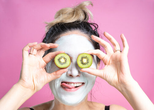 Portrait of a funny young woman in a cosmetic mask and kiwi instead of eyes on a pink background.