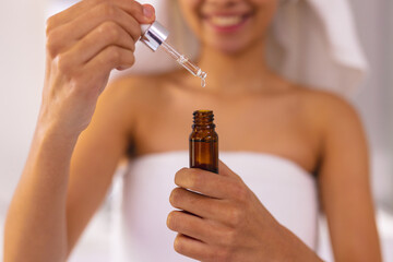Smiling biracial woman wearing towel in bathroom, holding essential oil dropper bottle, copy space
