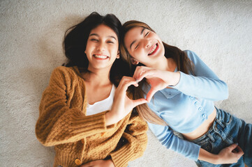 Top view two happy pretty asian women lying on carpet and posing together hand showing heart shape...