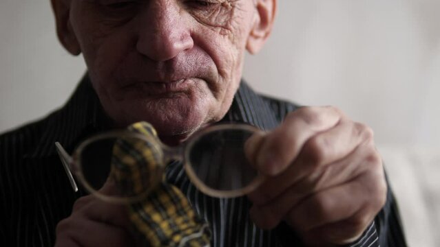  old man 70-79 years old wiping glasses from dirt and dust with a napkin. an elderly man cleans the lenses of glasses. loss of vision in old age. selective focus. lifestyle of an old man. 