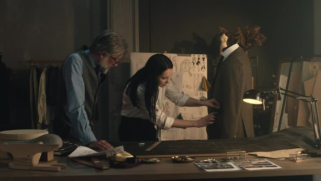 Male and female tailors working on sketch pattern of business or wedding suit. Mannequin with tailored shirt and jacket in stylish luxury designer atelier or tailoring dim studio. Fashion concept.