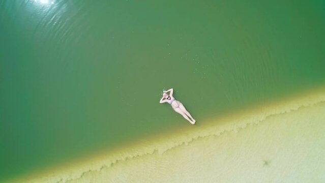 Young caucasian woman in sunglasses and swimsuit relaxes in water of salt lake Baskunchak. Aerial vertical shot.