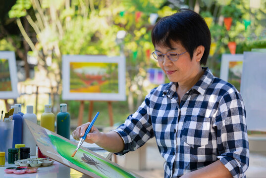 talented female artist sitting and holding paint brush painting watercolor on painting pad, middle aged asian woman having inspiration while working on a picture