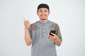 smiling or happy asian muslim man holding phone with thumbs up wearing grey muslim clothes isolated on white background