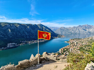 Montenegro flag blowing in wind with scenic view from St Ivan fortress on Kotor bay in sunny summer, Adriatic Mediterranean Sea, Montenegro, Balkan Peninsula, Europe. Fjord winding along coastal towns