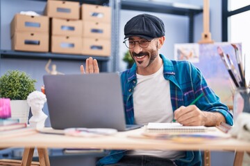 Hispanic man with beard doing online video call at art studio looking positive and happy standing and smiling with a confident smile showing teeth