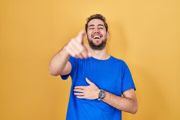 Hispanic man with beard standing over yellow background laughing at you, pointing finger to the camera with hand over body, shame expression