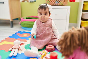 Adorable hispanic baby sitting on floor holding toy at kindergarten