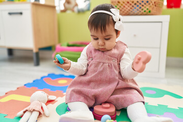 Adorable hispanic baby sitting on floor holding toys at kindergarten