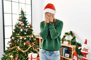 Redhead man with long beard wearing christmas hat by christmas tree with sad expression covering face with hands while crying. depression concept.