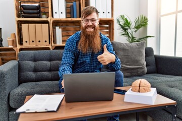 Redhead man with long beard using laptop working at psychology clinic smiling happy and positive,...