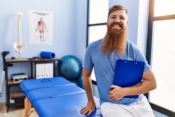 Fototapeta premium Young redhead man wearing physiotherapist uniform holding clipboard at physiotherapy clinic