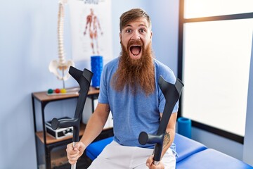 Redhead man with long beard holding crutches at rehabilitation clinic celebrating crazy and amazed...