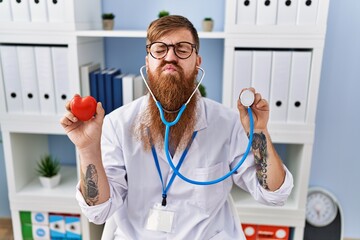 Redhead man with long beard wearing doctor uniform holding heart and stethoscope looking at the...
