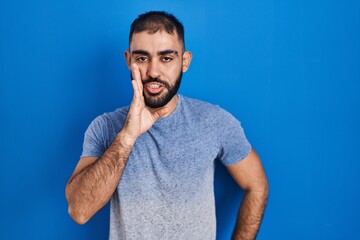 Middle east man with beard standing over blue background hand on mouth telling secret rumor, whispering malicious talk conversation