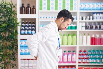 Handsome hispanic man working at pharmacy drugstore suffering of backache, touching back with hand,...