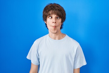 Hispanic young man standing over blue background making fish face with lips, crazy and comical gesture. funny expression.