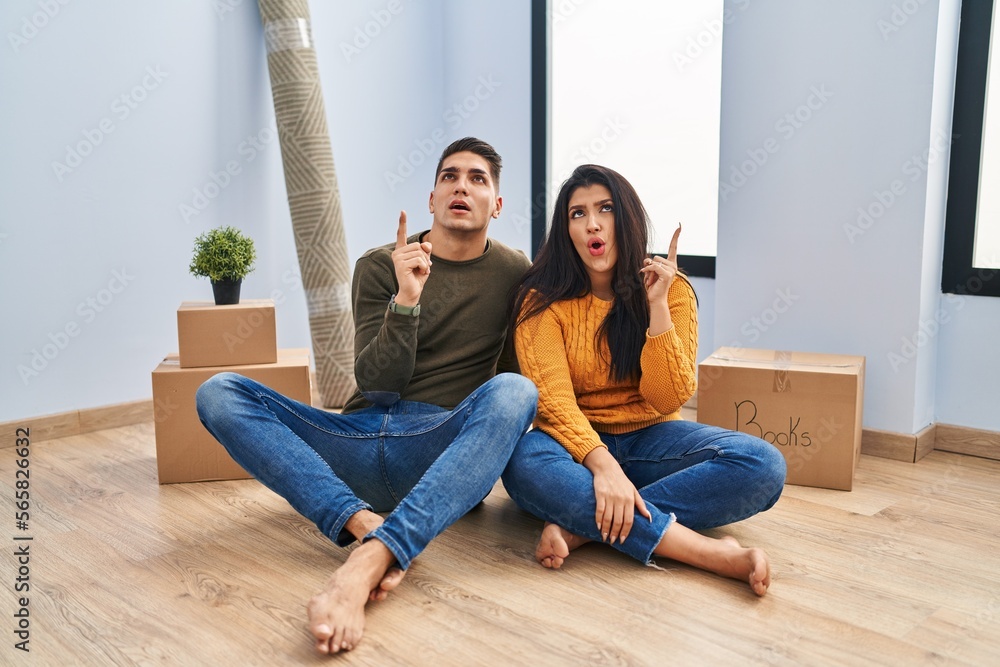 Poster Young couple sitting on the floor at new home amazed and surprised looking up and pointing with fingers and raised arms.