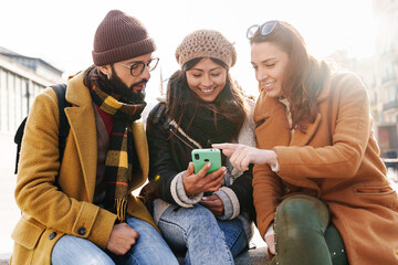 Group of friends sitting together using mobile phone to share content on social media in winter 