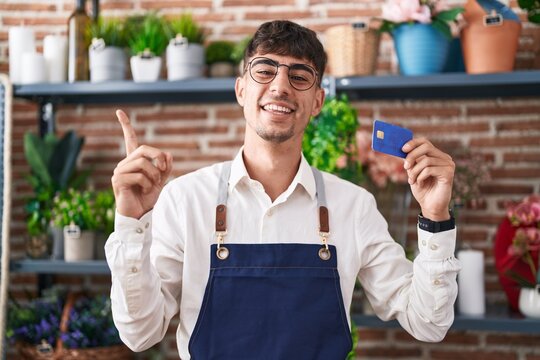 Young hispanic man working at florist shop holding credit card smiling happy pointing with hand and finger to the side