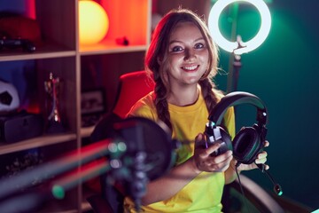 Young blonde woman streamer smiling confident holding headphones at gaming room